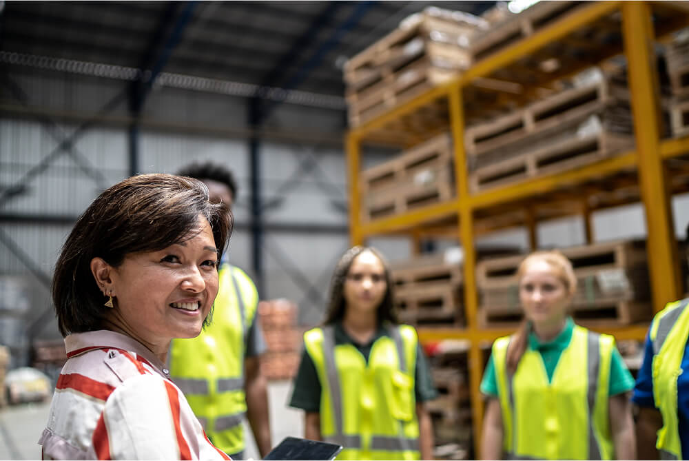 A photo of a group of workers in a warehouse setting standing in front of their team leader. She is smiling to camera.