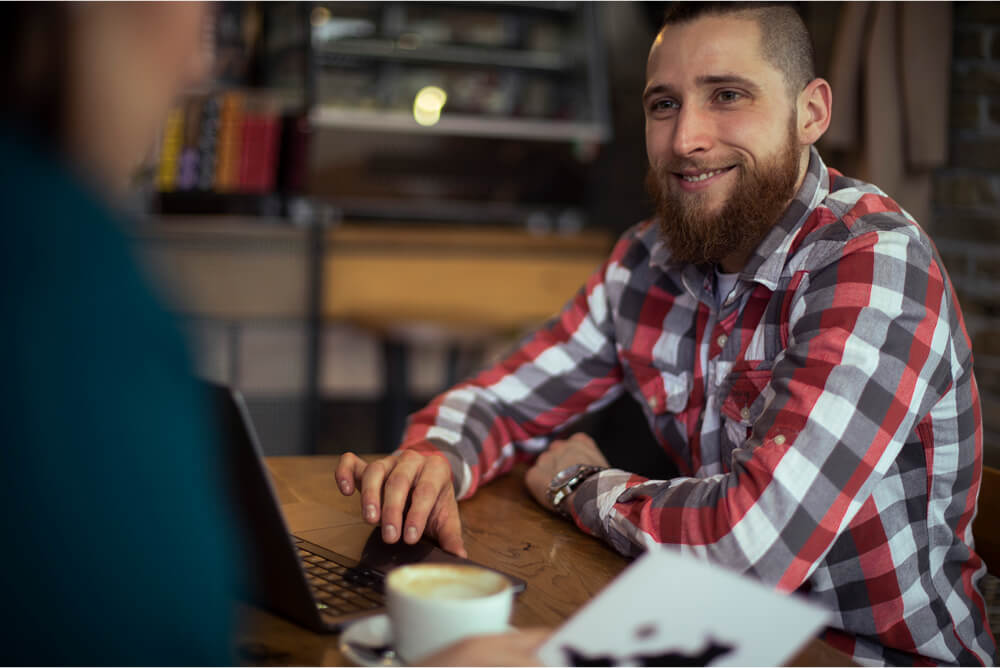 A photo of a man with a beard in a cafe sitting opposite another person. He is smiling.