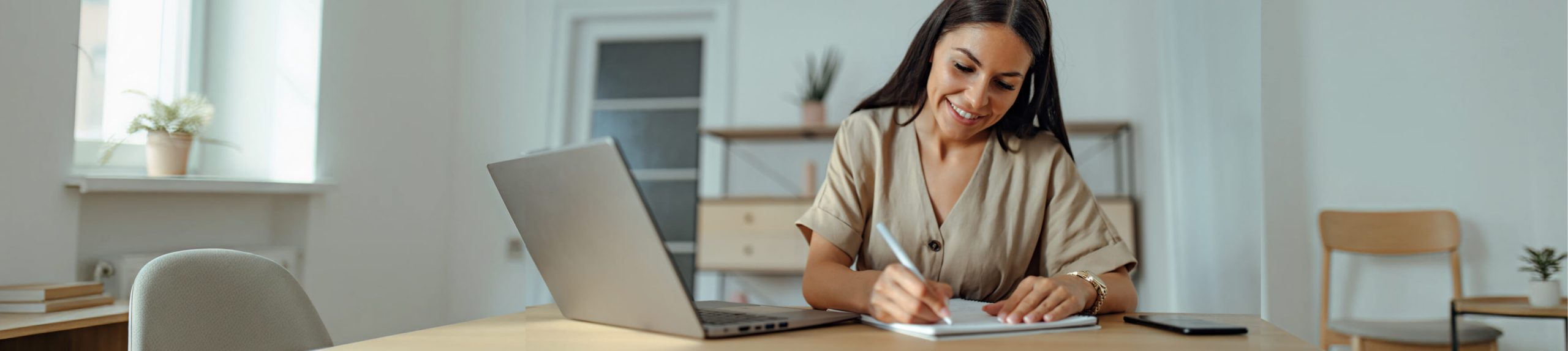 A woman sitting writing on a pad in front of her laptop.